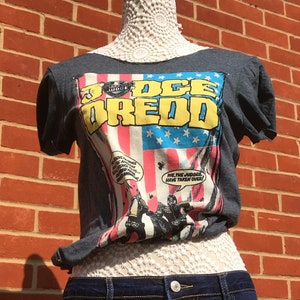 K&ouml;nnte beinhalten: Dunkelgraues T-Shirt mit Comic-Grafik mit den Worten "Judge Dredd" in Gelb und Blau. Das Shirt wird mit zweifarbigen Jeansshorts kombiniert. Die Schaufensterpuppe steht vor einer roten Backsteinmauer.