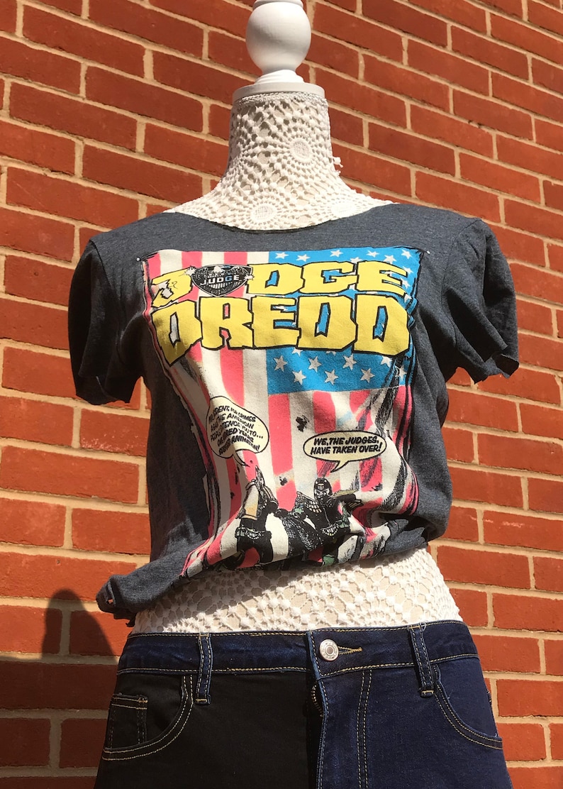 K&ouml;nnte beinhalten: Dunkelgraues T-Shirt mit Comic-Grafik mit den Worten "Judge Dredd" in Gelb und Blau. Das Shirt wird mit zweifarbigen Jeansshorts kombiniert. Die Schaufensterpuppe steht vor einer roten Backsteinmauer.