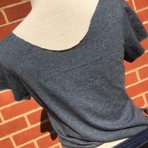 K&ouml;nnte beinhalten: Ein meliertes graues T-Shirt mit tiefem R&uuml;ckenausschnitt und kurzen &Auml;rmeln. Das Shirt wird auf einer Schaufensterpuppe pr&auml;sentiert, wobei der R&uuml;cken und der Ausschnitt sichtbar sind. Das Shirt wird mit einer dunkelblauen Jeans kombiniert.