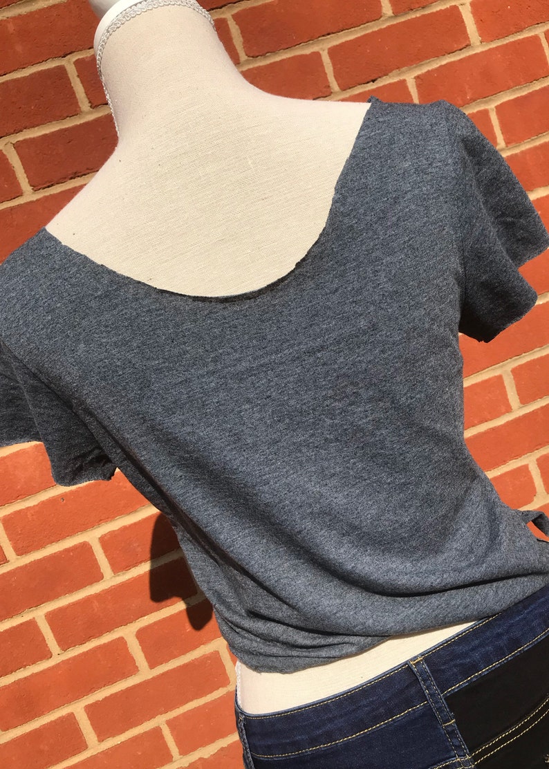 K&ouml;nnte beinhalten: Ein meliertes graues T-Shirt mit tiefem R&uuml;ckenausschnitt und kurzen &Auml;rmeln. Das Shirt wird auf einer Schaufensterpuppe pr&auml;sentiert, wobei der R&uuml;cken und der Ausschnitt sichtbar sind. Das Shirt wird mit einer dunkelblauen Jeans kombiniert.