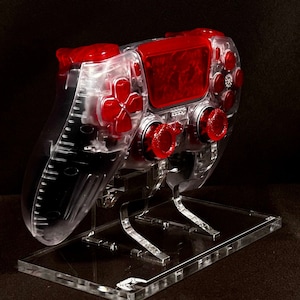 May include: A clear, translucent PlayStation 4 controller with red buttons and a red faceplate. The controller is displayed on a clear acrylic stand.
