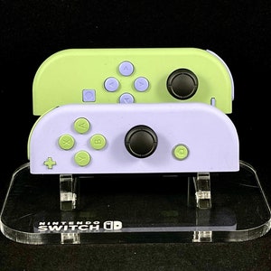 May include: Two pastel-colored Nintendo Switch Joy-Cons, one light green and one lavender, are stacked on a clear acrylic stand. The stand is labeled "NINTENDO SWITCH" in white text. The Joy-Cons have black control sticks and colored buttons.