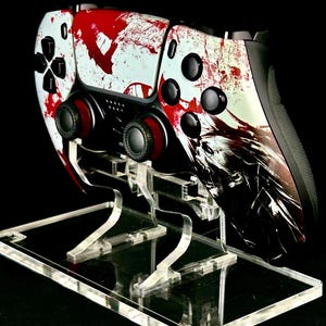 Peut inclure: Une manette de jeu vidéo personnalisée avec un motif éclaboussé blanc et rouge, posée sur un support en acrylique transparent. La manette a des boutons noirs et des joysticks rouges. Le support est rectangulaire avec des supports incurvés.