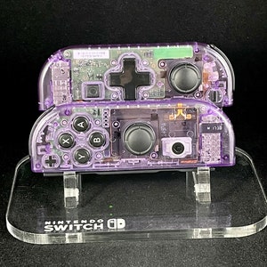 May include: Two clear purple Nintendo Switch Joy-Con controllers on a clear acrylic stand. The controllers are disassembled, showing the internal components.