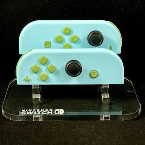 May include: Two light blue Nintendo Switch controllers with green buttons and a black analog stick, displayed on a clear plastic stand with the text "Nintendo Switch" printed on the front.