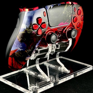 Peut inclure: Une manette de jeu vidéo personnalisée, rouge et noire, posée sur un présentoir en acrylique transparent. La manette présente un graphisme détaillé et des boutons rouges. Le présentoir est rectangulaire avec plusieurs supports.