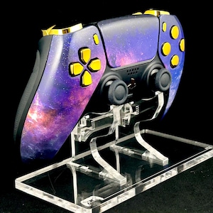 Puede incluir: Un mando de PlayStation 5 con temática de galaxia con botones dorados y un soporte de acrílico transparente. El mando tiene un diseño de galaxia morado y azul con detalles dorados.
