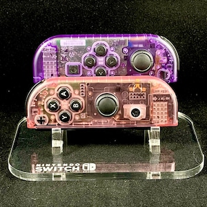 May include: Two clear plastic Nintendo Switch Joy-Con controllers, one purple and one pink, are displayed on a clear acrylic stand. The stand has the words "Nintendo Switch" printed on it.