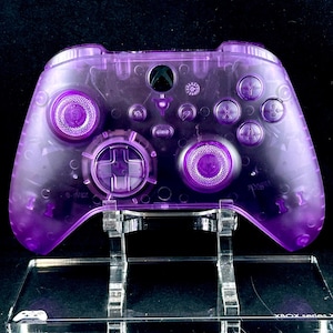 Op de afbeelding: Een transparante paarse Xbox Series X-controller op een transparante acrylstandaard. De controller heeft een transparante paarse behuizing en paarse knoppen.