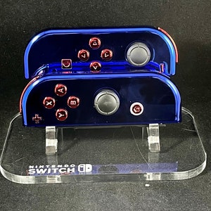 May include: Two blue Nintendo Switch Joy-Con controllers with red buttons are displayed on a clear acrylic stand. The stand has the words "Nintendo Switch" printed on it.