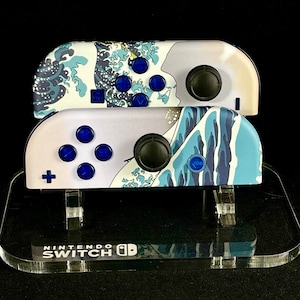 Könnte beinhalten: Zwei blaue und weiße Nintendo Switch Joy-Con-Controller mit Wellendesign auf einem durchsichtigen Acryl-Ständer. Der Ständer trägt den Schriftzug "Nintendo Switch".