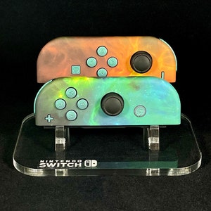May include: Two Nintendo Switch Joy-Con controllers with custom galaxy-themed designs. The top controller has a fiery orange and brown design, while the bottom controller has a blue, green, and purple galaxy design. The controllers are displayed on a clear acrylic stand with the Nintendo Switch logo.