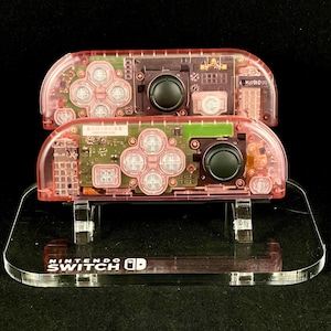 Könnte beinhalten: Zwei rosa Nintendo Switch-Controller auf einem durchsichtigen Acryl-Ständer mit dem Aufdruck "Nintendo Switch" auf der Basis.