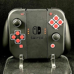 May include: A black Nintendo Switch console with a clear acrylic stand. The console has red buttons and the text "NINTENDO SWITCH" on the front.