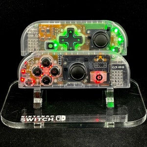 May include: Two clear plastic Nintendo Switch controllers with red and green lights on the buttons. The controllers are on a clear plastic stand with the words "Nintendo Switch" printed on it.