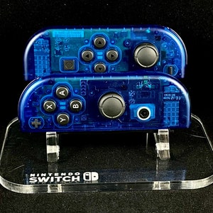 May include: Two transparent blue Nintendo Switch Joy-Con controllers on a clear acrylic stand with the text "NINTENDO SWITCH" printed on the base.