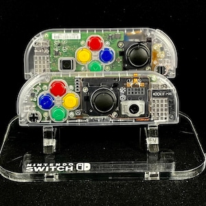 May include: Two clear plastic Nintendo Switch Joy-Cons with colorful buttons and a clear plastic stand with the text "NINTENDO SWITCH" printed on it.