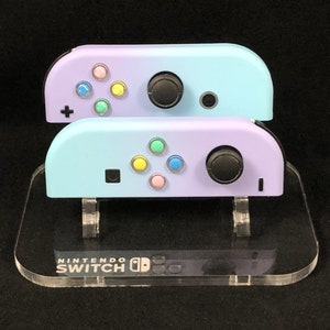 May include: Two pastel-colored Nintendo Switch Joy-Con controllers on a clear acrylic stand with the Nintendo Switch logo. The top controller is light blue with a purple hue, and the bottom controller is light blue.