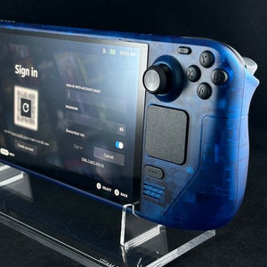 Könnte beinhalten: Eine blaue Steam Deck Handheld-Gaming-Konsole mit einem durchsichtigen Kunststoffgehäuse wird auf einem durchsichtigen Acrylständer präsentiert. Die Konsole zeigt einen Anmeldebildschirm mit dem Text "Sign in" und einem QR-Code.