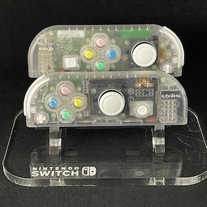 May include: Two clear plastic Nintendo Switch Joy-Con controllers on a clear plastic stand. The stand has the words "Nintendo Switch" printed on it.