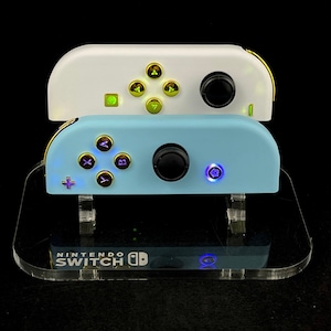 Puede incluir: Dos mandos de Nintendo Switch en un soporte de acrílico transparente. El mando superior es blanco con botones verdes y detalles dorados. El mando inferior es azul claro con botones morados y detalles dorados. El soporte tiene el logotipo de Nintendo Switch impreso.
