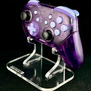 May include: A transparent purple Nintendo Switch Pro controller on a clear acrylic stand. The controller has black and light purple buttons. The stand has the Nintendo Switch logo.