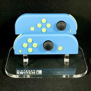 May include: Two blue Nintendo Switch controllers with light green buttons and a clear acrylic stand. The stand has the text "NINTENDO SWITCH" printed on it.