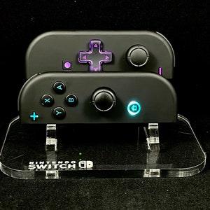 May include: Two black Nintendo Switch controllers with purple and blue lights on the buttons. The controllers are on a clear acrylic stand with the text "Nintendo Switch" printed on it.