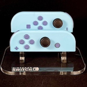 May include: Two light blue Nintendo Switch Joy-Cons with purple buttons are displayed on a clear acrylic stand. The stand has the Nintendo Switch logo printed on it.
