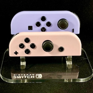 Puede incluir: Dos mandos Joy-Con de Nintendo Switch en un soporte de acrílico transparente. El mando superior es lavanda y el mando inferior es rosa.