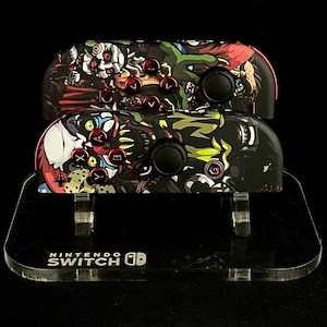 May include: Two black and red Nintendo Switch Joy-Con controllers with colorful, graphic designs featuring monsters and clowns. The controllers are displayed on a clear acrylic stand with the text "NINTENDO SWITCH" printed on the front.