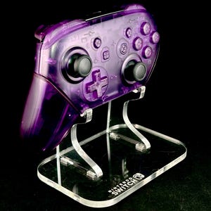 Op de afbeelding: Een transparante paarse Nintendo Switch Pro Controller staat op een heldere acryl standaard. De controller heeft zwarte joysticks en knoppen. De standaard heeft het Nintendo Switch logo op de basis, tegen een zwarte achtergrond.