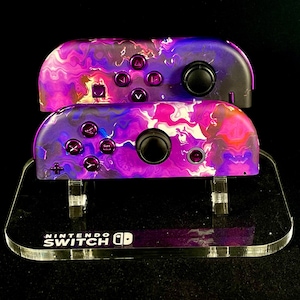 May include: Two purple and black Nintendo Switch Joy-Con controllers with a marbled design, displayed on a clear acrylic stand with the text "NINTENDO SWITCH" printed on the base.
