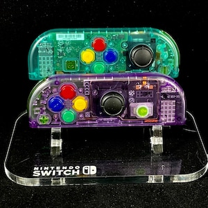 May include: Two clear plastic Nintendo Switch Joy-Con controllers, one purple and one teal, with colorful buttons, are displayed on a clear plastic stand with the words "Nintendo Switch" printed on it.