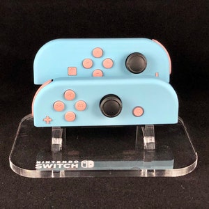 May include: Two light blue Nintendo Switch Joy-Con controllers with pink buttons are displayed on a clear acrylic stand. The stand has the Nintendo Switch logo printed on the front.