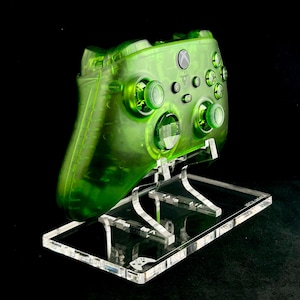 May include: A translucent green Xbox controller on a clear acrylic stand. The controller has a green button and a green Xbox logo.