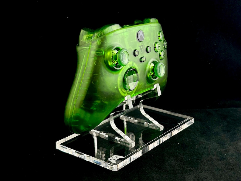May include: A translucent green Xbox controller on a clear acrylic stand. The controller has a green button and a green Xbox logo.