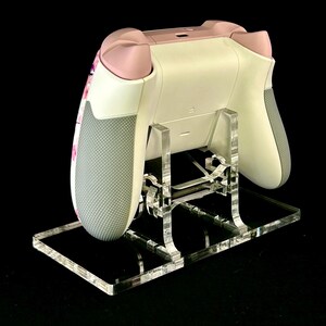 May include: A white and pink video game controller rests on a clear acrylic stand. The controller has gray grips and pink accents. The stand is made of clear acrylic and has a rectangular base.
