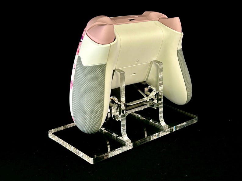 May include: A white and pink video game controller rests on a clear acrylic stand. The controller has gray grips and pink accents. The stand is made of clear acrylic and has a rectangular base.