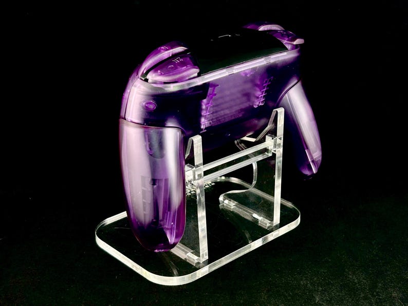 Op de afbeelding: Een transparante paarse gamecontroller op een heldere acryl standaard. De controller heeft een glanzende afwerking en is ontworpen voor gaming. De standaard is rechthoekig met een gebogen rand en ondersteunt de controller.