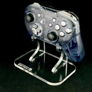 May include: A transparent Nintendo Switch Pro controller on a clear acrylic stand. The controller has black buttons and a silver directional pad. The stand has the Nintendo Switch logo on the base. The background is black.
