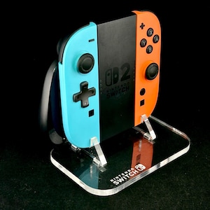 Könnte beinhalten: Eine Nintendo Switch-Konsole mit blauen und orangefarbenen Joy-Con-Controllern wird auf einem transparenten Acryl-Ständer präsentiert. Der Ständer trägt das Nintendo Switch-Logo. Die Controller sind schwarz, blau und orange.