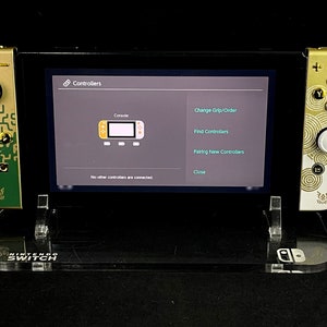 K&ouml;nnte beinhalten: Eine goldene und gr&uuml;ne Nintendo Switch-Konsole mit benutzerdefinierten Joy-Cons, die auf einem klaren Acryl-St&auml;nder pr&auml;sentiert wird. Der Bildschirm der Konsole zeigt das Men&uuml; "Controller" mit Optionen zum &Auml;ndern der Griffreihenfolge, zum Finden von Controllern, zum Koppeln neuer Controller und zum Schlie&szlig;en.