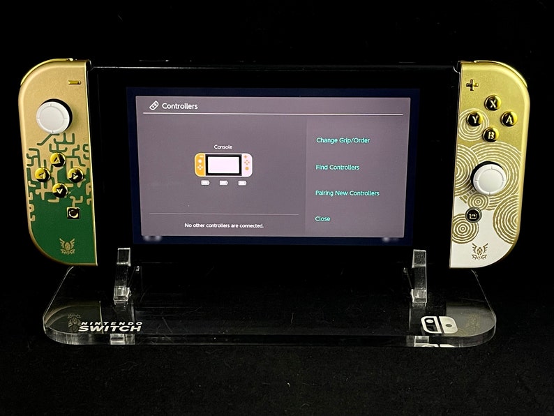 K&ouml;nnte beinhalten: Eine goldene und gr&uuml;ne Nintendo Switch-Konsole mit benutzerdefinierten Joy-Cons, die auf einem klaren Acryl-St&auml;nder pr&auml;sentiert wird. Der Bildschirm der Konsole zeigt das Men&uuml; "Controller" mit Optionen zum &Auml;ndern der Griffreihenfolge, zum Finden von Controllern, zum Koppeln neuer Controller und zum Schlie&szlig;en.