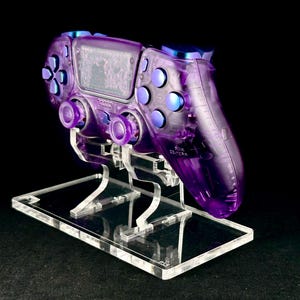 May include: A transparent purple games controller with blue highlights, displayed on a clear acrylic stand. The controller has buttons and joysticks, and the stand has a rectangular base.