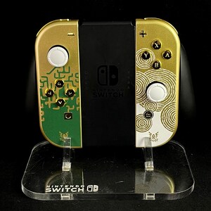 K&ouml;nnte beinhalten: Eine goldene Nintendo Switch-Konsole mit einem gr&uuml;n-goldenen Design auf dem linken Joy-Con und einem wei&szlig;-goldenen Design auf dem rechten Joy-Con. Die Konsole steht auf einem durchsichtigen Acryl-St&auml;nder mit dem Nintendo Switch-Logo.