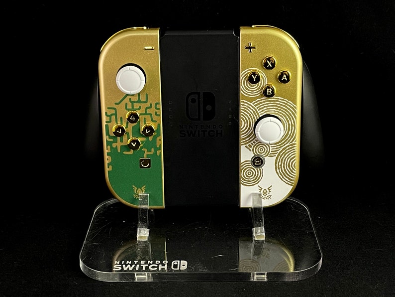 K&ouml;nnte beinhalten: Eine goldene Nintendo Switch-Konsole mit einem gr&uuml;n-goldenen Design auf dem linken Joy-Con und einem wei&szlig;-goldenen Design auf dem rechten Joy-Con. Die Konsole steht auf einem durchsichtigen Acryl-St&auml;nder mit dem Nintendo Switch-Logo.