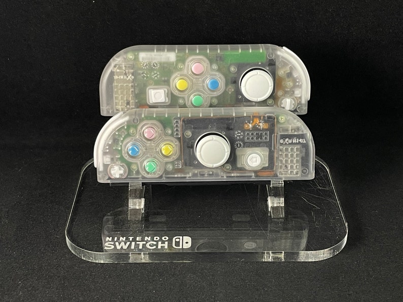 May include: Two clear plastic Nintendo Switch Joy-Con controllers on a clear plastic stand. The stand has the words "Nintendo Switch" printed on it.