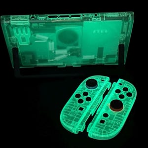 May include: Glow-in-the-dark transparent green gaming console and controllers. The console is displayed on a black stand, with two detached controllers below. The controllers have black and brown buttons. The internal components are visible through the transparent casing.