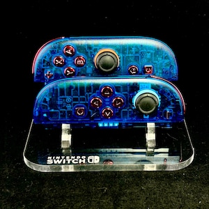 Könnte beinhalten: Zwei transparente blaue Nintendo Switch Joy-Con-Controller mit roten Knöpfen und schwarzen Joysticks sind auf einem transparenten Acrylständer gestapelt. Der Ständer hat das Nintendo Switch-Logo. Die Controller sind von innen beleuchtet.